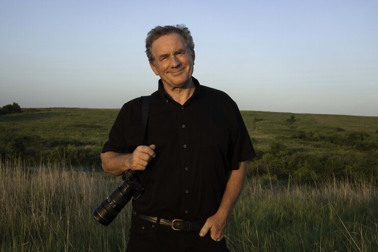 Joel Sartore stands in a field, camera hanging from right shoulder