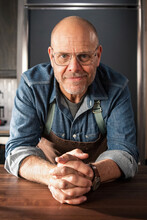 Alton Brown, wearing a chef's apron, leans over a counter, with his hands crossed in front of him.