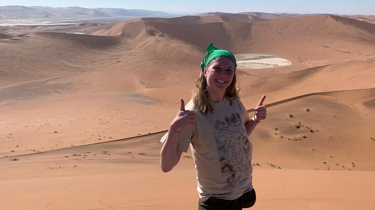 A young woman stands amid sand dunes, smiling and giving two thumbs-up.