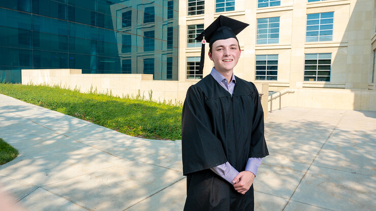 Business student Caleb Newburn stands in his graduation gown outside of the College of Business' Hawks Hall.