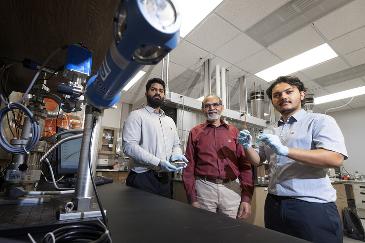 Abhijeet Prasad (left), a doctoral student in engineering; Ravi Saraf (center), Anderson Distinguished Professor of chemical and biomolecular engineering; and Aashish Subedi, a senior physics major, are photographed in Saraf’s Othmer Hall lab. Prasad is holding small electronic chips used to study the nanoparticle necklace network devices. Subedi is holding a suspension of nanoparticle necklaces (blue container) made from individual particles (red container).