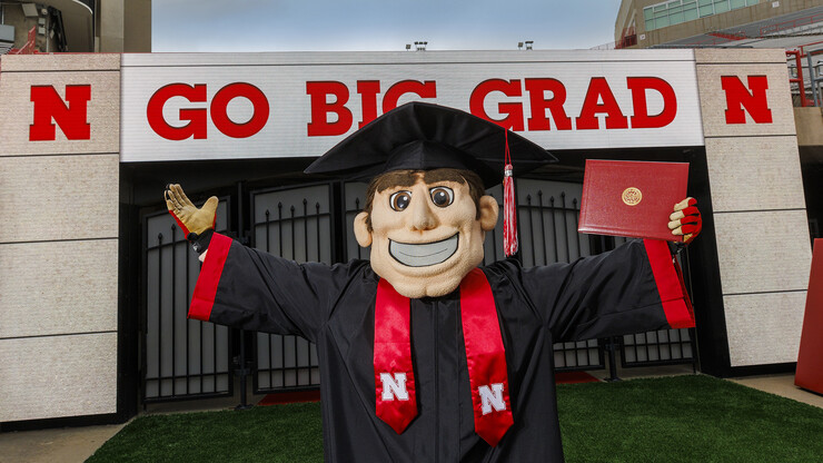 Herbie Husker, in graduation attire, holds a diploma on the field at Memorial Stadium