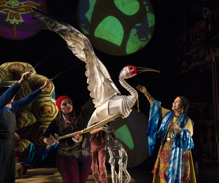 Two puppeteers control a crane puppet next to woman in colorful garments