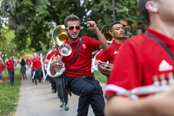 Cornhusker Marching Band members in red shirts and black pants march down the sidewalk near Kimball Hall