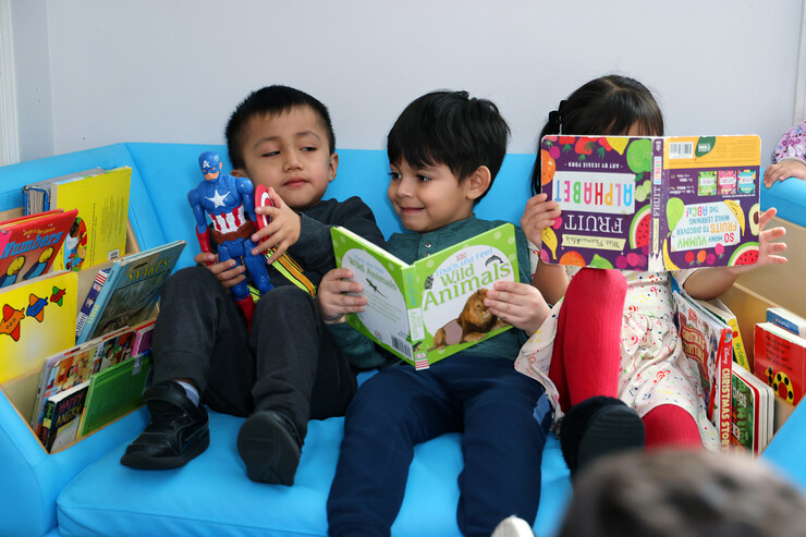 Three young children sit together, one holding a Captain America doll and two holding books