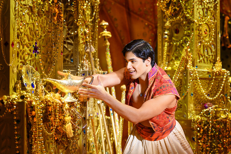 Adi Roy as Aladdin holds a golden lamp in a room full of treasure.