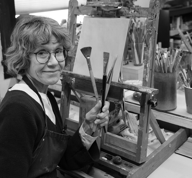 Black-and-white photo of Paula Wallace, holding up three paintbrushes in front of an easel.