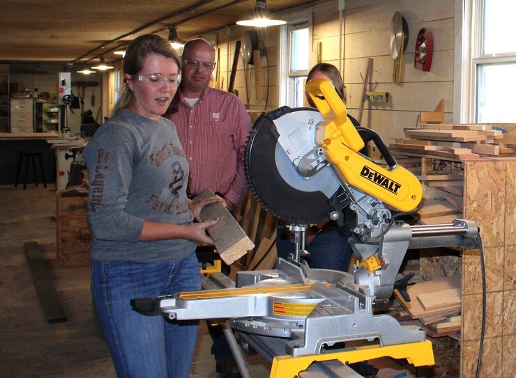 A young woman wearing safety goggles holds a board in front of a saw as a man and woman watch.