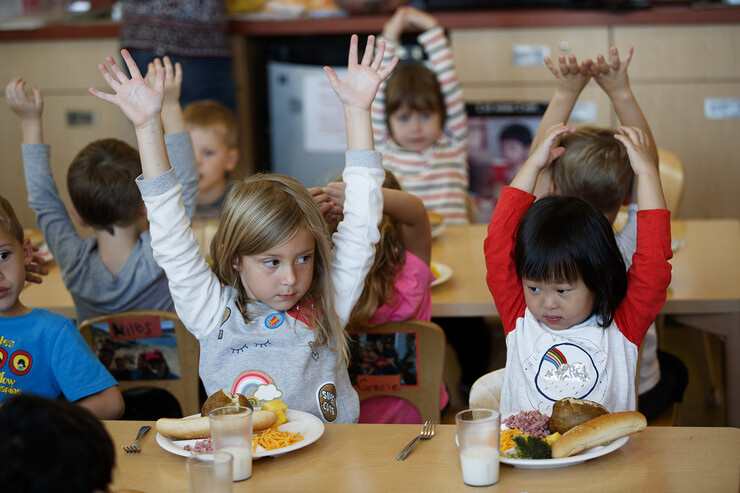 Young children raise their hands in a lunchroom.