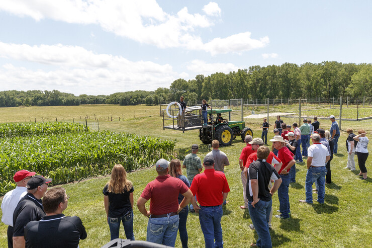 About two dozen people stand near a hail simulator and a corn plot.