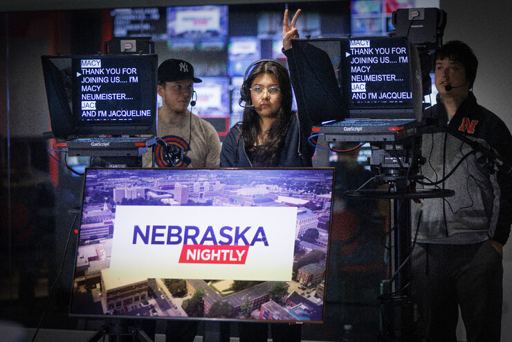 A young woman and two young men stand behind two teleprompters and a screen that reads "Nebraska Nightly."
