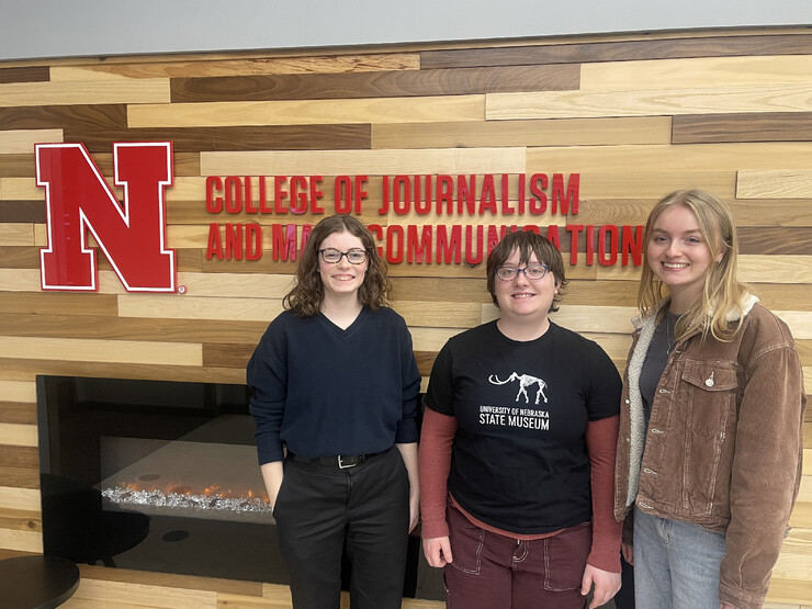 Husker journalists Shelby Rickert, Samantha Grove and Jordan Moore stand in front of an electric fireplace and lettering that read "College of Journalism and Mass Communications."
