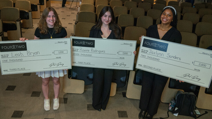 Three young women hold giant checks in an auditorium.