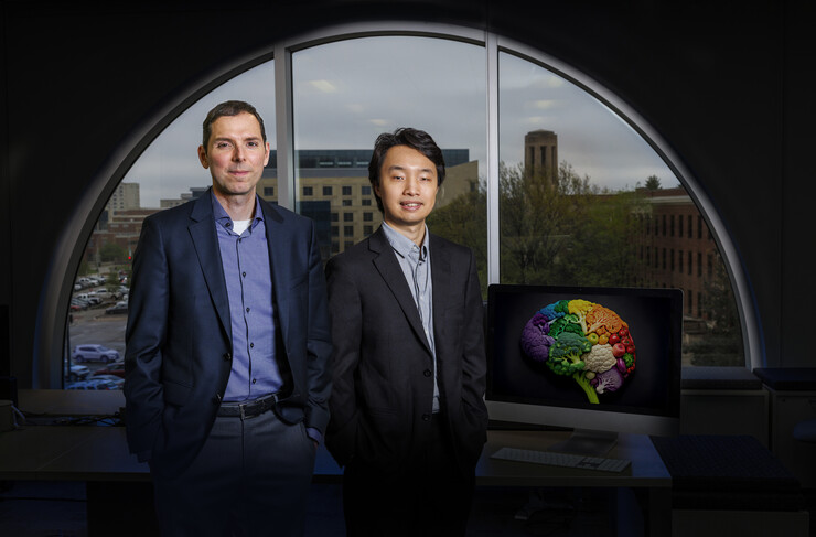 Aron Barbey and Jisheng Wu stand in front of a half-circle window, next to a monitor displaying a brain made of fruits and vegetables.