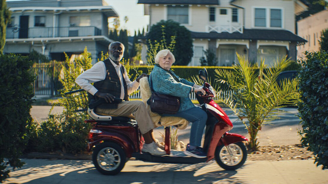 Scene from the film "Thelma" with two elderly people sitting on a motorized cart on a sidewalk in front of homes.