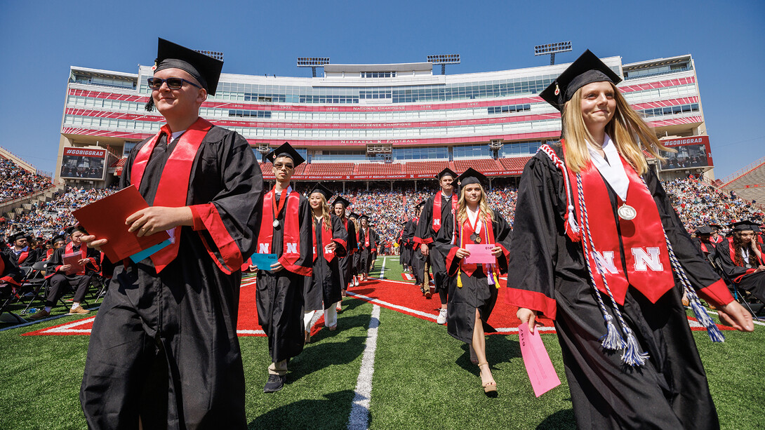 Husker graduates in commencement regalia walk in two lines at Memorial Stadium.