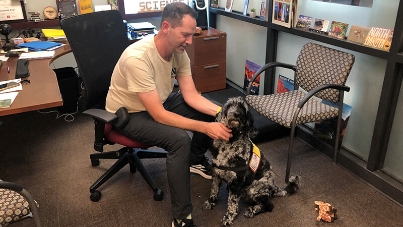 Kenneth Pyle sits in a black rolling office chair in a tan shirt and black jeans alongside River, a black and grey labradoodle.