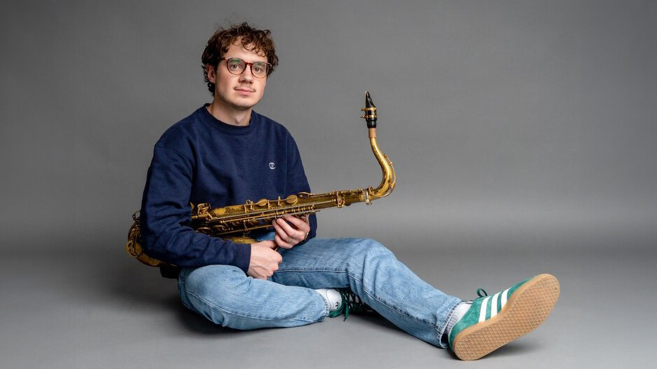 Pete Fucinaro sitting on the ground holding his saxophone and his right leg is extended outward.