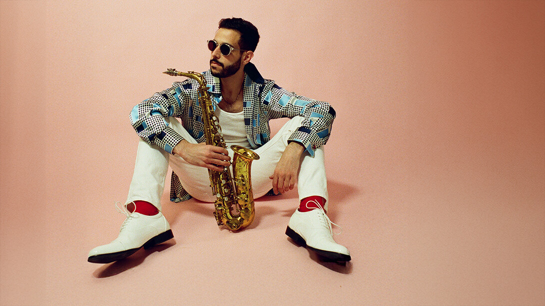 Performer Sam Greenfield sitting on a floor holding his saxophone.