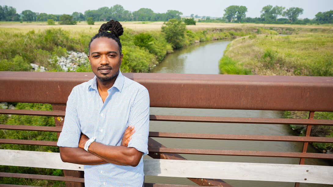 Ng’ang’a Wahu-Mũchiri, associate professor of English, stands on a bridge, over a river, with his arms crossed. He is wearing a blue-and-white plaid shirt.
