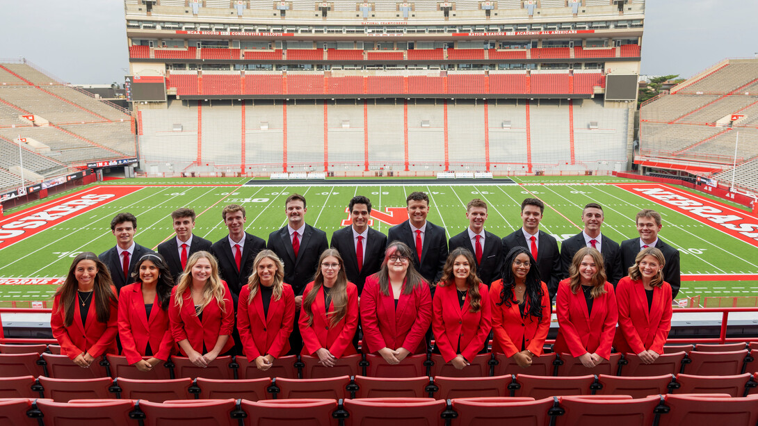 Ten young men and 10 young women — all 2025 homecoming royalty finalists — pose for a photo in the stands at Memorial Stadium, with the football field in the background. The men are wearing black suits with red ties, and the women are wearing red blazers with black shirts.