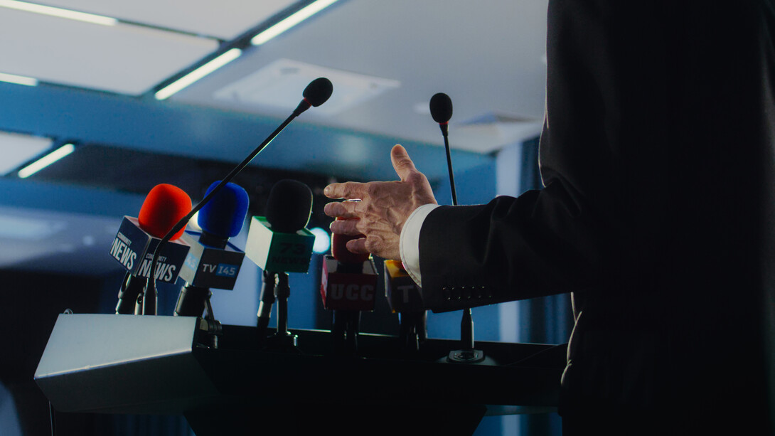 A man in a black suit gestures behind a lectern filled with microphones.