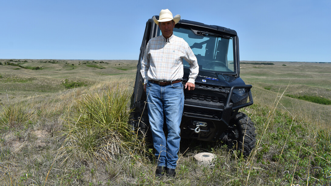 Homel Buell, owner and operator of Shovel Dot Ranch near Rose, Nebraska, stands in front of an all-terrain vehicle in a field. He is wearing a light-colored longsleeve plaid shirt, blue jeans and a white cowboy hat.