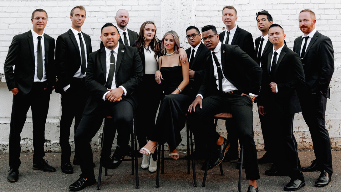 Ten men and two women wearing black and white formal attire pose for a photo in front of a white brick wall.