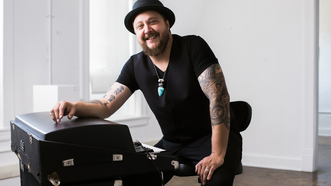 Josh Hoyer, wearing a black V-neck and black hat, sits next to an electric organ.