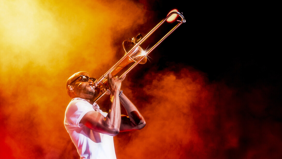 Musician Trombone Shorty, wearing a white short-sleeve shirt and white pants, plays a trombone on a smoke-filled stage.