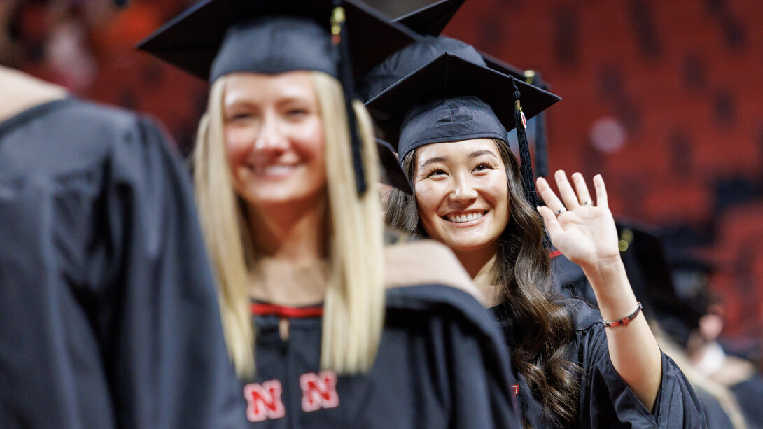 A female graduate smiles as another female graduate smiles and waves in line at Pinnacle Bank Arena.