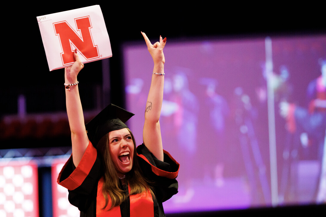 A female graduate throws her hands in the air in excitement as she crosses the stage at Pinnacle Bank Arena. She holds her newly earned diploma in her right hand. 