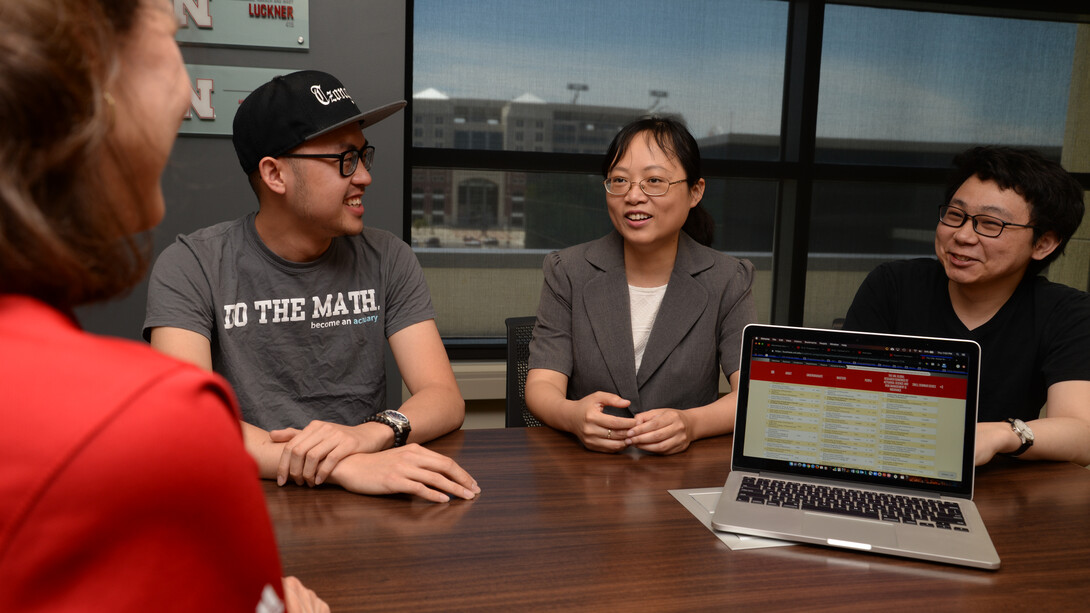 Two men and two women sit around a table, with an open laptop on it. Memorial Stadium can be seen through a window in the background.