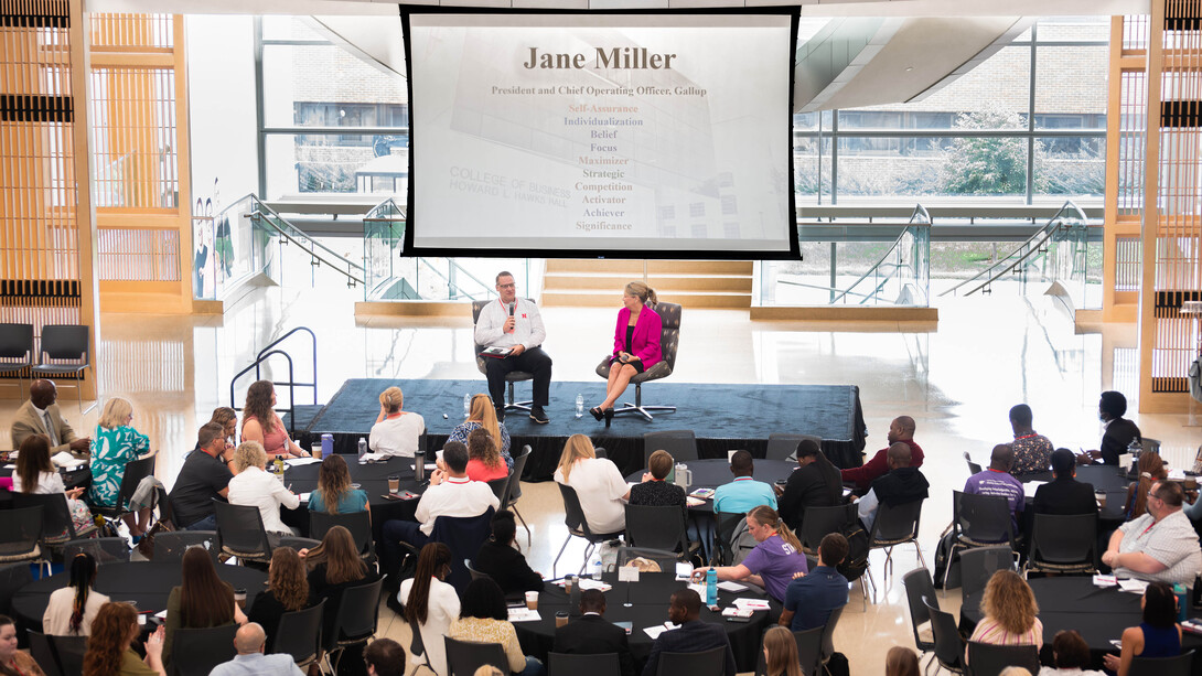 A crowd sits at tables around a podium, where a man in a white button-up shirt and black slacks interviews a woman in a black dress with a bright pink blazer. A projector presenting above the two show that the woman is Jane Miller.