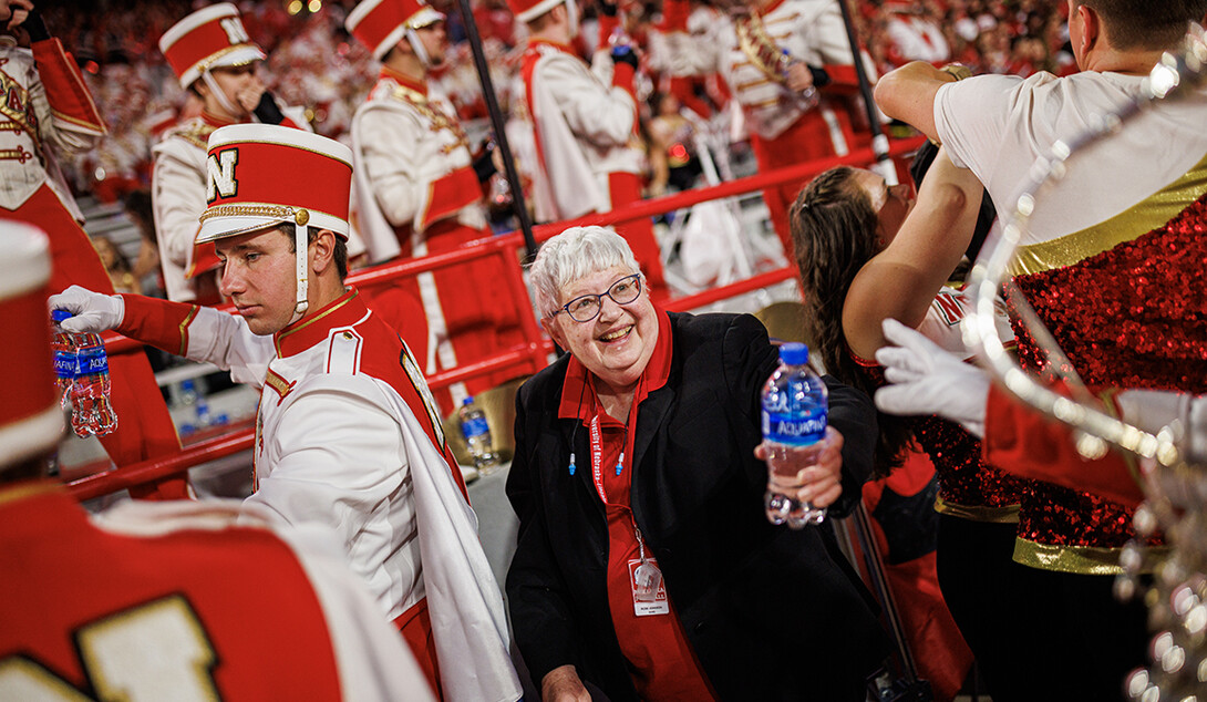 Rose Johnson stands in a crowd of marching band students wearing a red collared shirt and black blazer, with short gray hair and glasses.
