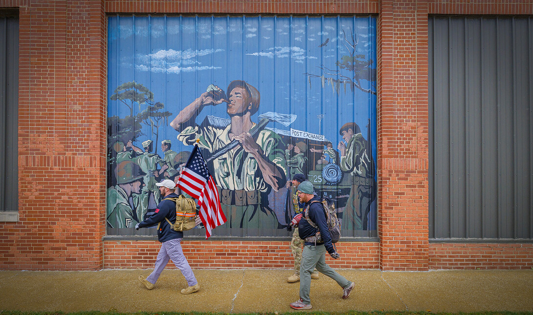 Nebraska alumni and veteran Pete Lass carries the American flag as he and Atlantic-area veterans Trevor Gipple (carrying game ball) and Ryan Graham walk past a mural on the side of the Coca-Cola Bottling Company in downtown Atlantic, Iowa, on the second day of the march.