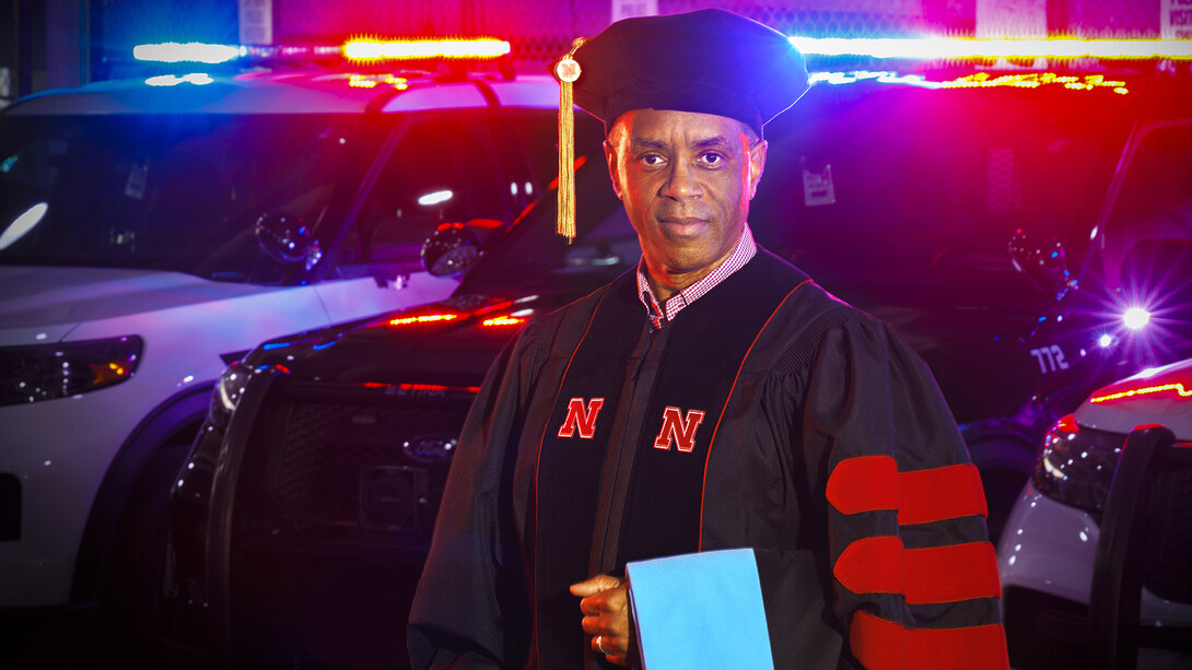 UNL Police Chief Hassan Ramzah standing in doctoral regalia with police cars in the background with their red and blue lights shining.