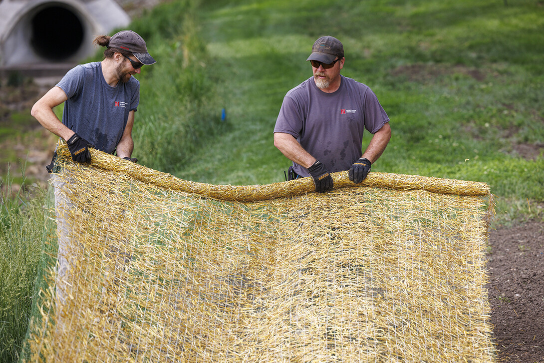 Ben Holmes, left, and Brian Dieterman unroll a straw mat to cover newly seeded ground around the new pedestrian bridge at East Campus’ Maxwell Arboretum.