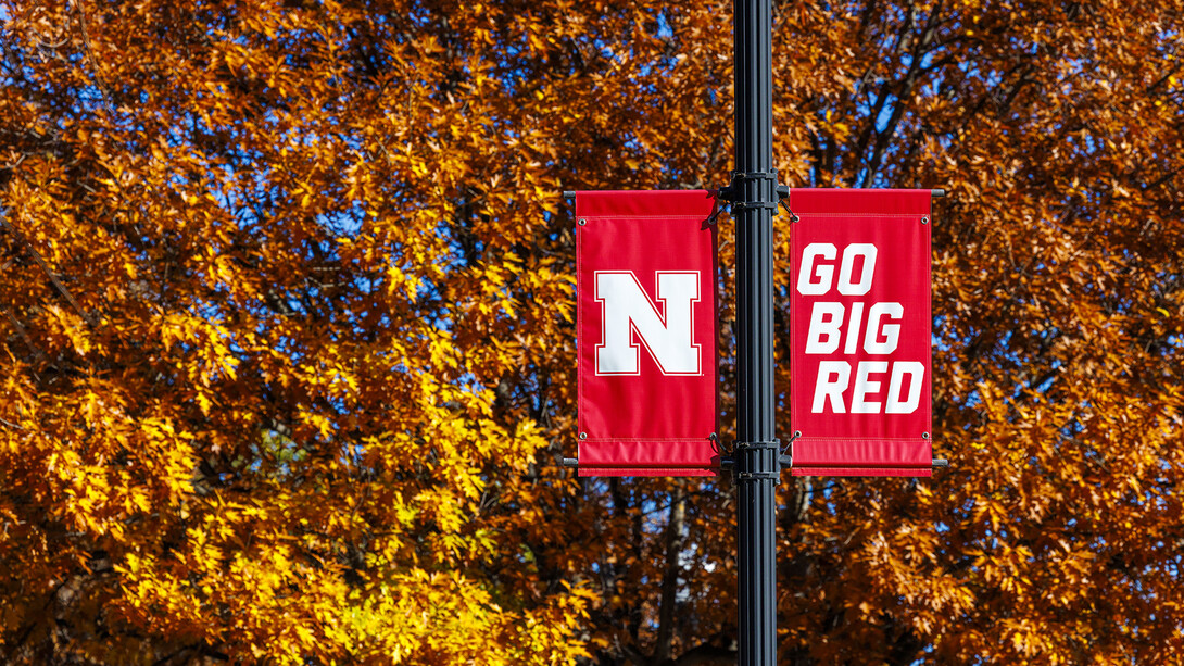 Nebraska "Go Big Red" banners on a street pole in front of yellow and orange fall leaves on a tree.