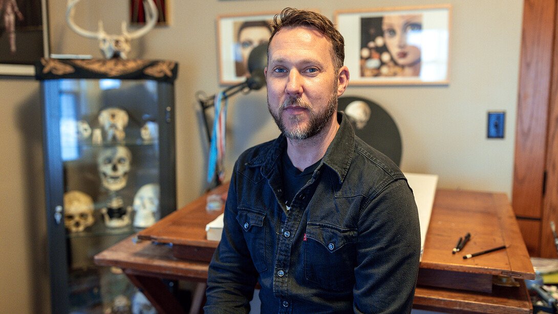 Scott McDonald sitting at his artist desk in his home studio with skulls in a cabinet in the background.