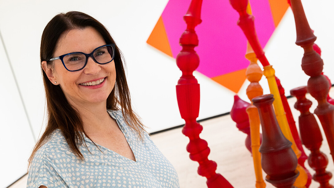 A white, brunette woman with black glasses and a light blue blourse stands next to a sculpture of multiple red, yellow and orange oddly-shaped tubes.