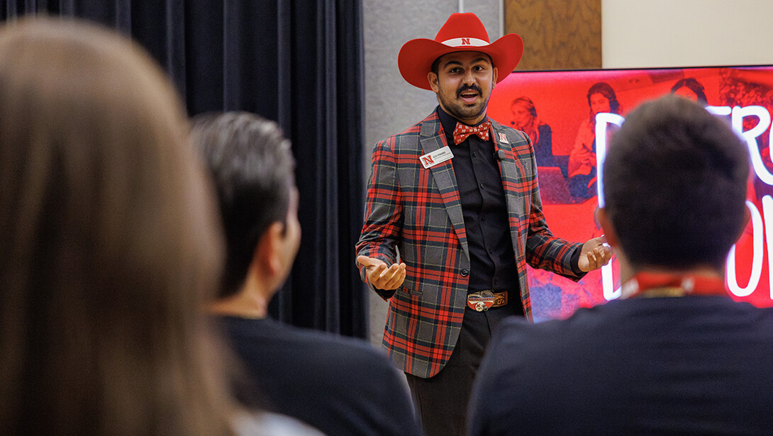 A man with brown skin, dressed in a black button-up shirt, black and red plaid blazer, red bowtie, and red cowboy hat presents to parents of prospective Huskers.