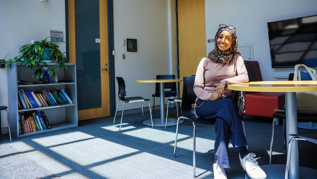 A young woman in a head covering sits at a table in a student workspace with a book shelf and computer screen behind her.