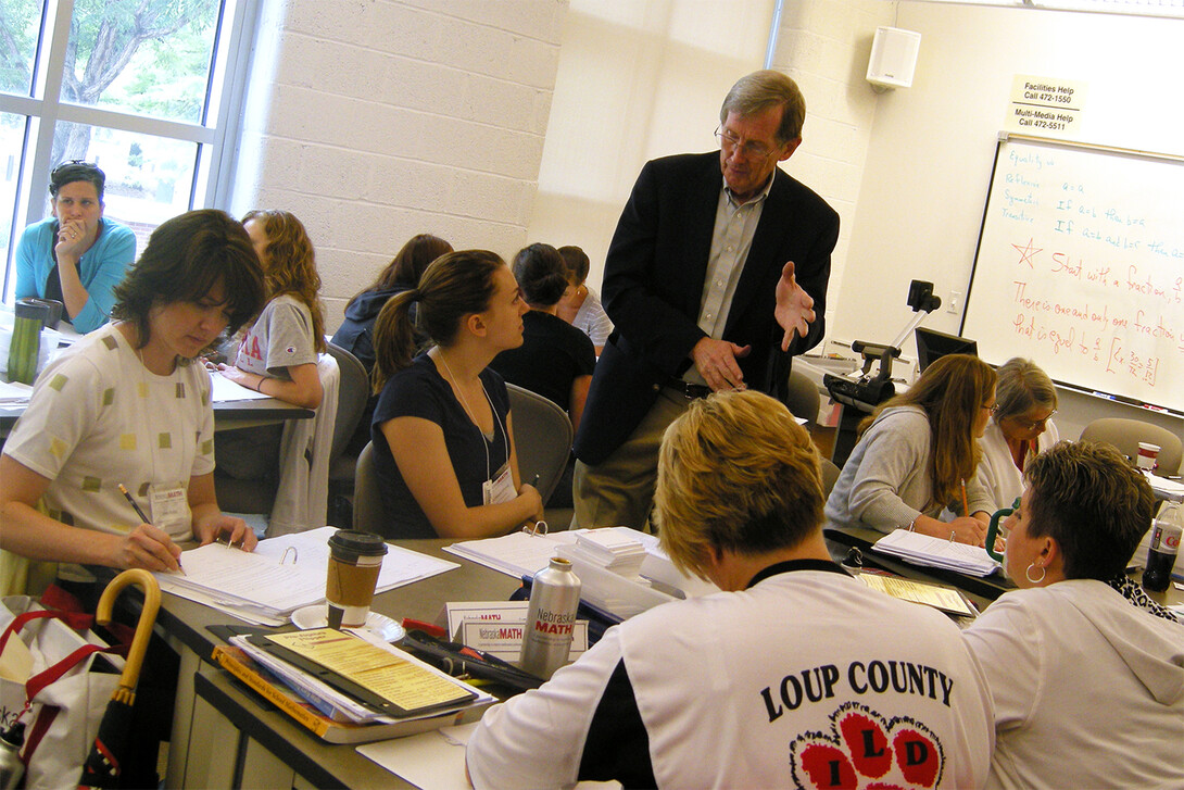 Jim Lewis stands in a classroom and speaks with teachers seated around a table
