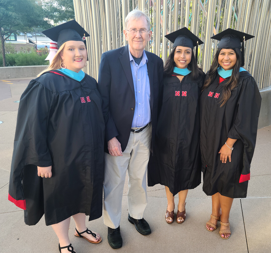 Jim Lewis stands with three master's graduates dressed in cap and gown