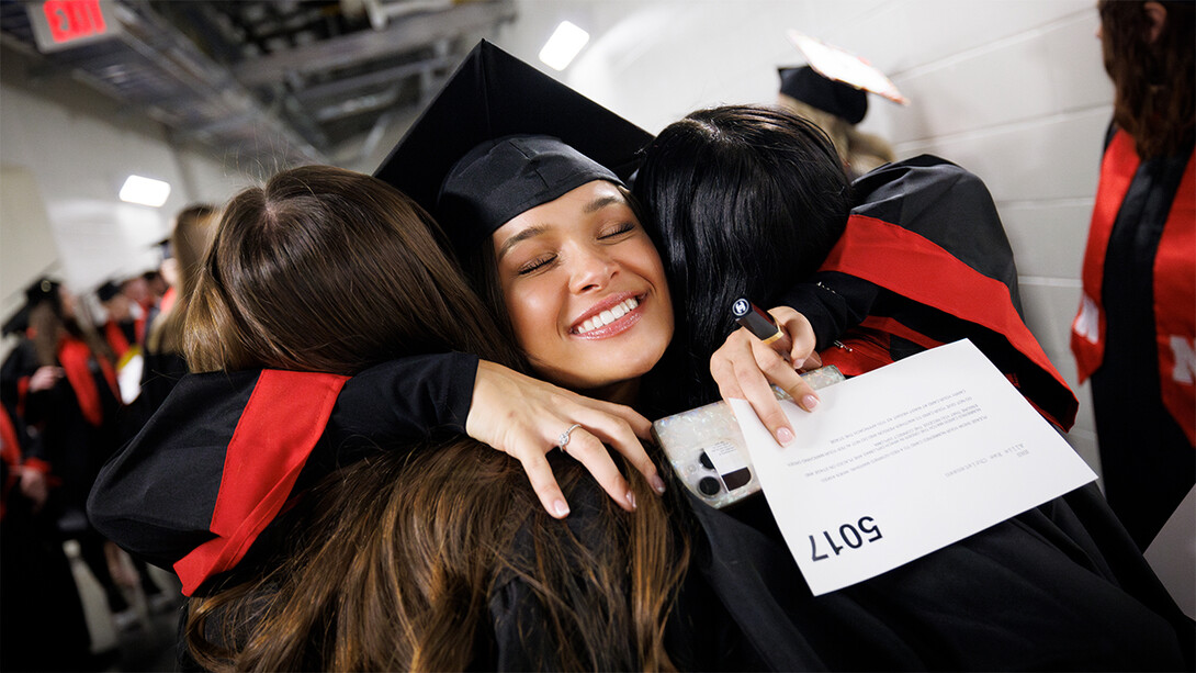 Allie Christensen, decked out in cap and gown, smiles with closed eyes while hugging two friends