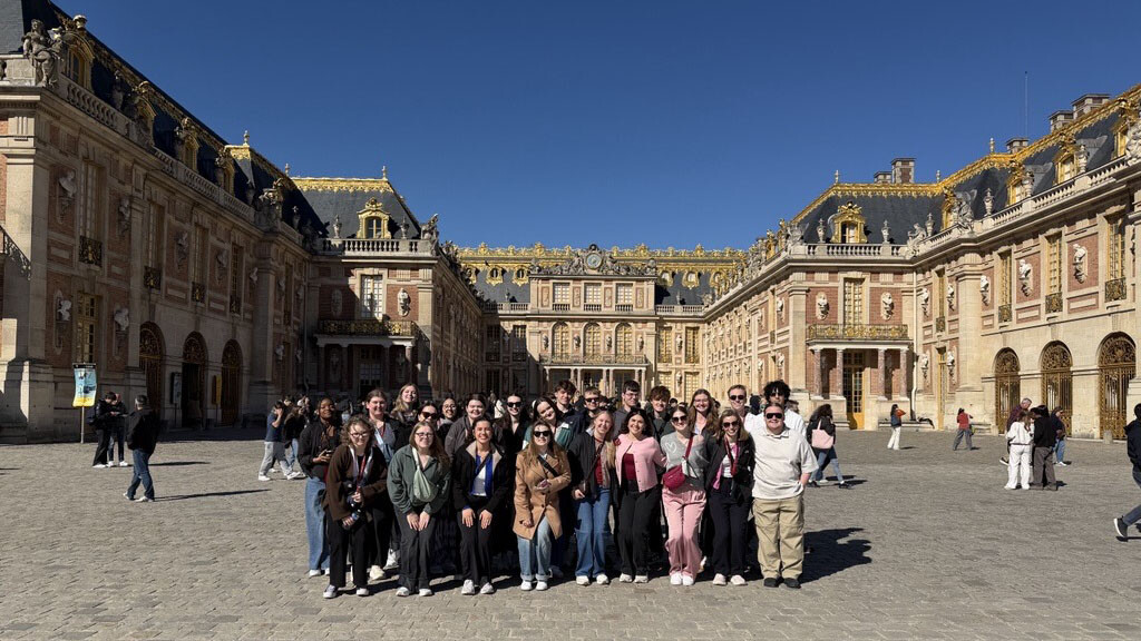 A large group of 25 students gathers in a group photo in front of a grand, stone building with a dark roof that forms a square around them in Paris.
