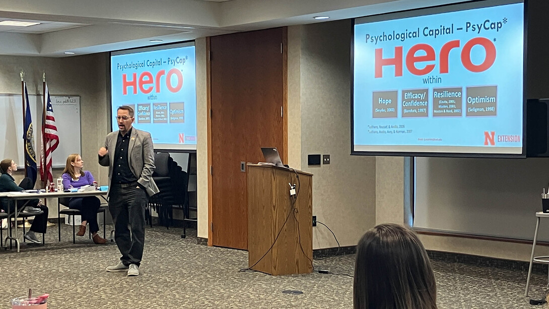 A brunette man in glasses, a black sweater and jeans and a grey blazer presents in front of two projectors displaying information on the PsyCap HERO program.