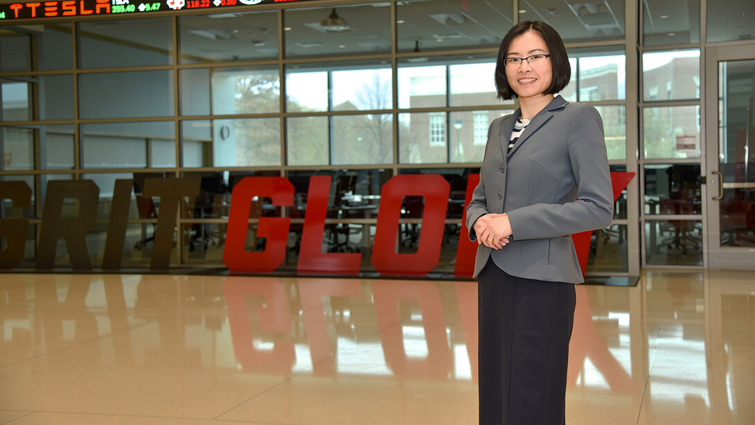 A brunette woman with a bob stands with her hands together, wearing a grey blazer, black slacks and thin-framed glasses.