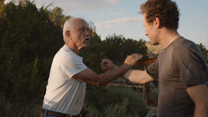 Patrick McCollum, a tan-skinned older man with grey hair and a mustache stands in a white t-shirt teaching Gabe Polsky, a white man with brown curly hair standing in a brown t-shirt, how to do a fighting move.